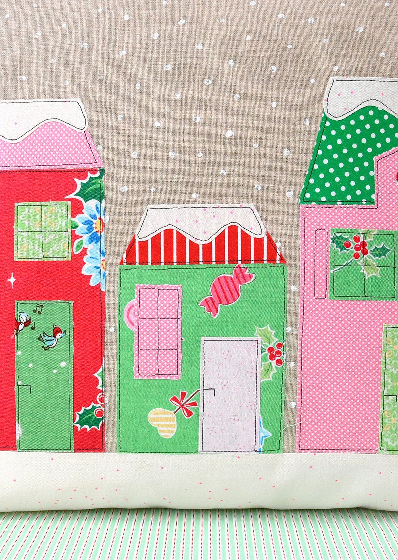May include: A festive pillow with a snowy scene featuring three houses decorated with red, green, and pink fabrics. The houses have windows, doors, and candy cane accents. The background is a beige fabric with white polka dots.