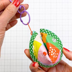 May include: A fabric fortune cookie keyring featuring a green polka dot border and a multi-coloured patchwork centre with orange, pink, yellow, and white patterns. The pouch includes a purple plastic lobster clasp and a small orange zip pull.
