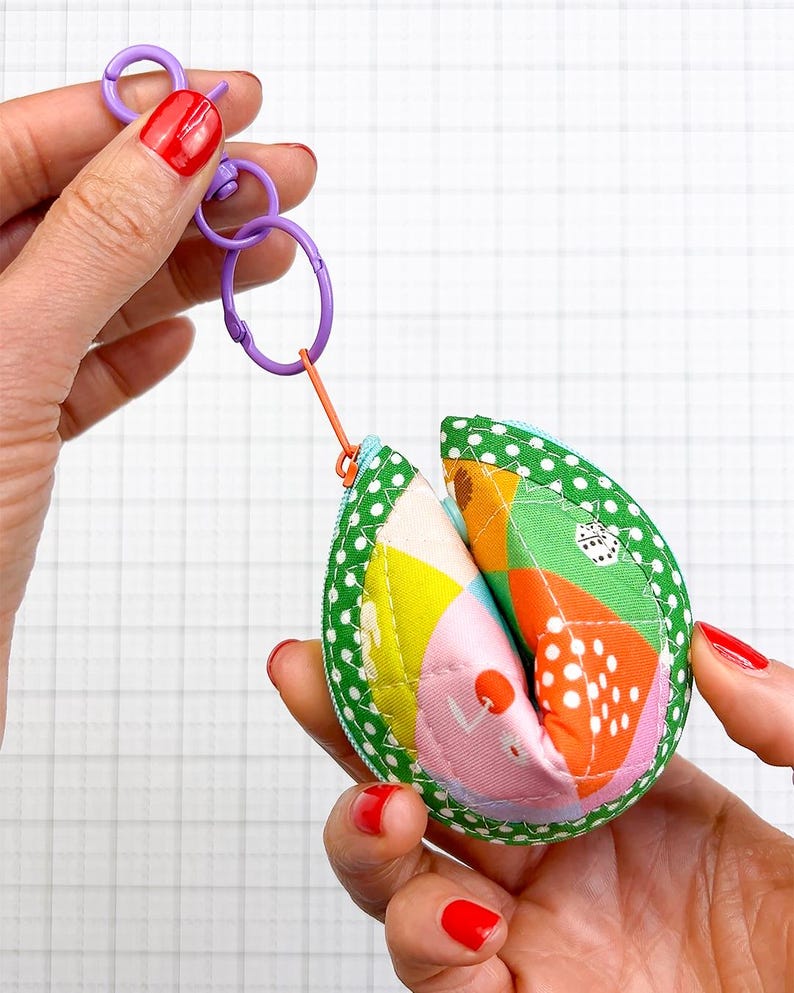 May include: A fabric fortune cookie keyring featuring a green polka dot border and a multi-coloured patchwork centre with orange, pink, yellow, and white patterns. The pouch includes a purple plastic lobster clasp and a small orange zip pull.