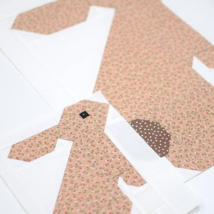 K&ouml;nnte beinhalten: Zwei Stoff-Hasen-Quiltbl&ouml;cke. Die Hasen sind aus beigem Stoff mit einem kleinen rosa Blumenmuster gefertigt. Das Auge des Hasen ist ein kleines schwarzes Quadrat. Der Schwanz des Hasen ist ein braunes Tupfenmuster. Die Bl&ouml;cke befinden sich auf wei&szlig;em Hintergrund.