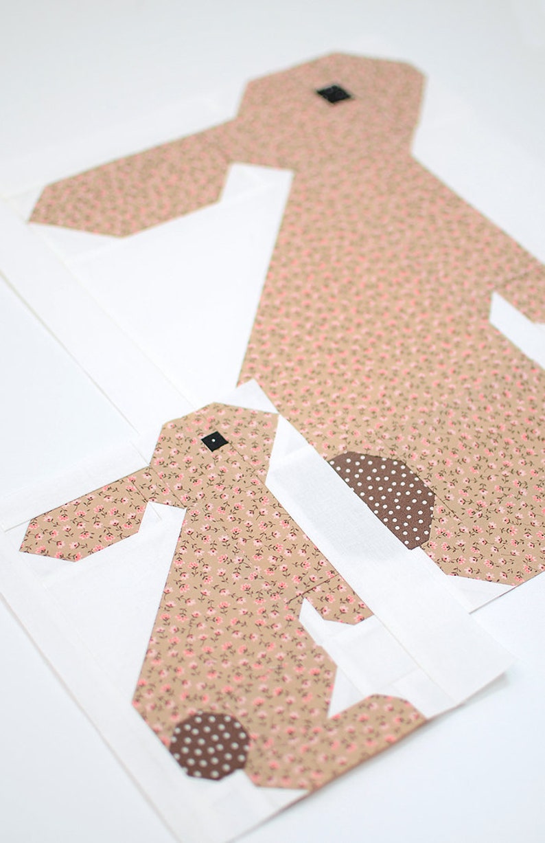 K&ouml;nnte beinhalten: Zwei Stoff-Hasen-Quiltbl&ouml;cke. Die Hasen sind aus beigem Stoff mit einem kleinen rosa Blumenmuster gefertigt. Das Auge des Hasen ist ein kleines schwarzes Quadrat. Der Schwanz des Hasen ist ein braunes Tupfenmuster. Die Bl&ouml;cke befinden sich auf wei&szlig;em Hintergrund.