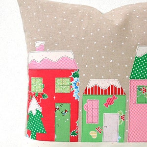 May include: A beige pillow with a snowy village scene. The houses are red, green, and pink with white trim and snow-covered roofs. The pillow has a white polka dot pattern.