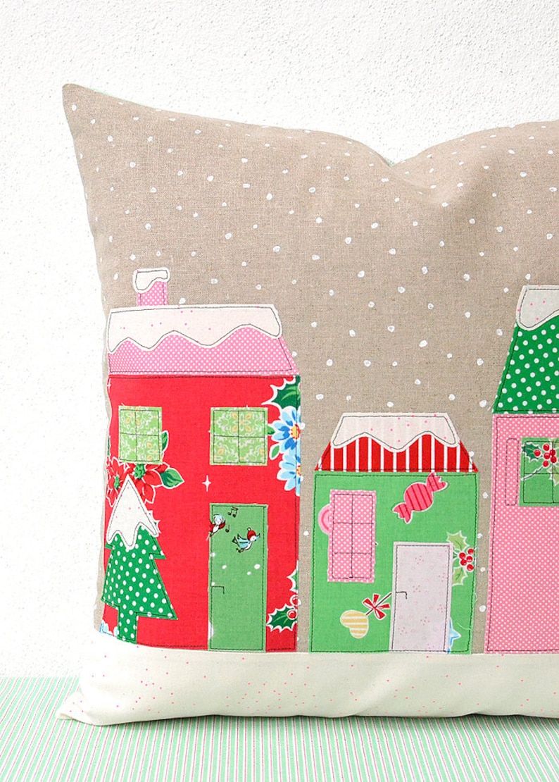 May include: A beige pillow with a snowy village scene. The houses are red, green, and pink with white trim and snow-covered roofs. The pillow has a white polka dot pattern.