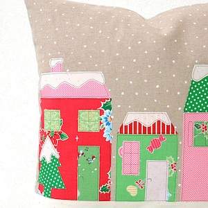 May include: A beige pillow with a snowy village scene. The houses are red, green, and pink with white trim and snow-covered roofs. The pillow has a white polka dot pattern.