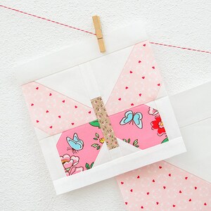 May include: A pink and white butterfly quilt block with a brown center and blue butterflies. The block is hanging from a red string with a wooden clothespin.