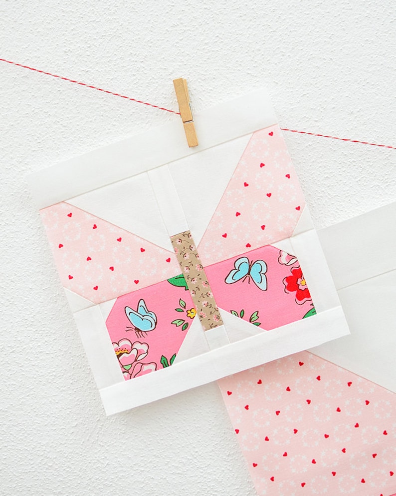 May include: A pink and white butterfly quilt block with a brown center and blue butterflies. The block is hanging from a red string with a wooden clothespin.