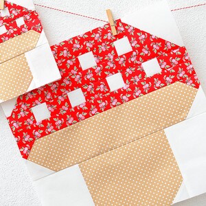 以下が含まれることがあります： 白い壁に赤い紐と木製の洗濯ばさみで吊るされた、赤と白のギンガムチェックのキノコのキルトブロック2つ。茶色の水玉模様の茎がついています。