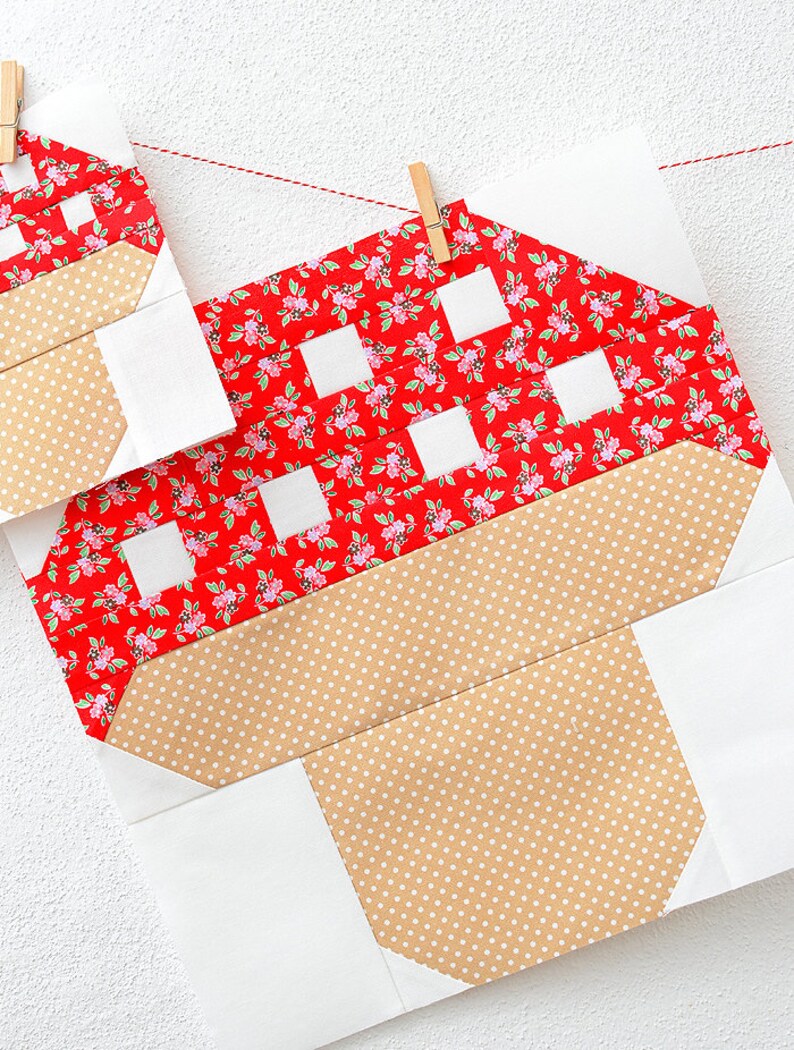 以下が含まれることがあります： 白い壁に赤い紐と木製の洗濯ばさみで吊るされた、赤と白のギンガムチェックのキノコのキルトブロック2つ。茶色の水玉模様の茎がついています。