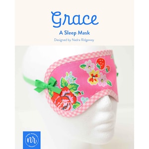 May include: A pink sleep mask featuring a floral pattern with red roses, yellow flowers, and strawberries. The mask has a pink and white gingham trim, a green ribbon bow on the side, and a green elastic strap. Text reads Grace A Sleep Mask.