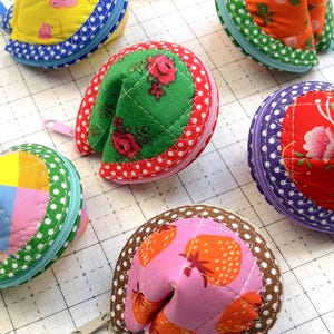 May include: A collection of six quilted fabric pouches shaped like fortune cookies, each featuring distinct patterns including yellow with pink pigs, orange with green leaves, green with red roses, purple with red flowers, and pink with strawberries.