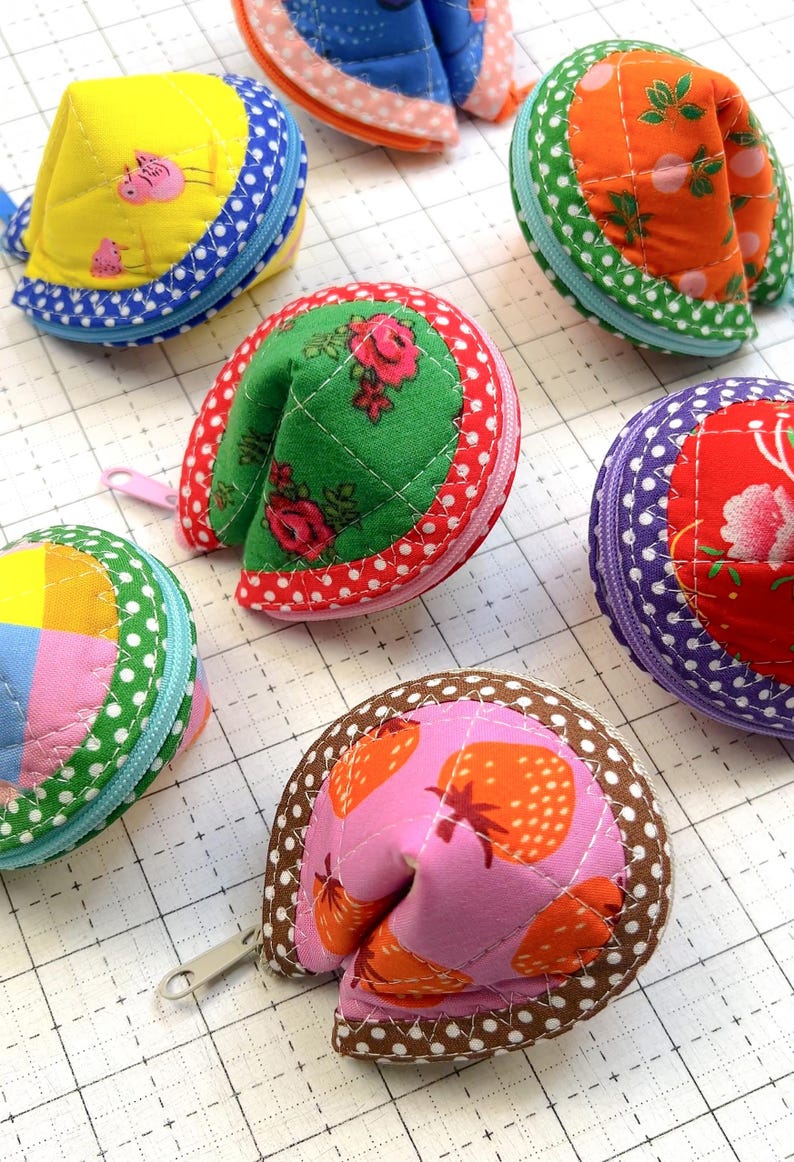 May include: A collection of six quilted fabric pouches shaped like fortune cookies, each featuring distinct patterns including yellow with pink pigs, orange with green leaves, green with red roses, purple with red flowers, and pink with strawberries.