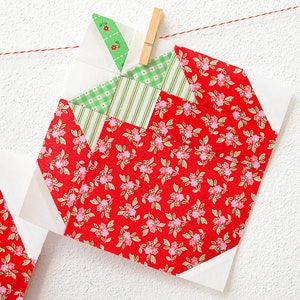 Peut inclure: Un bloc de courtepointe patchwork rouge, blanc et vert en forme de pomme. La pomme est faite de tissu &agrave; imprim&eacute; floral et est suspendue &agrave; une ficelle avec une pince &agrave; linge.