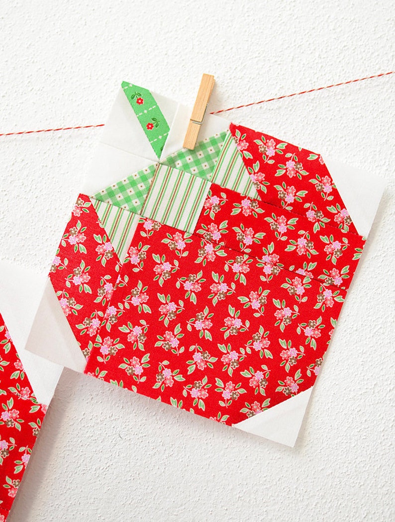 Peut inclure: Un bloc de courtepointe patchwork rouge, blanc et vert en forme de pomme. La pomme est faite de tissu &agrave; imprim&eacute; floral et est suspendue &agrave; une ficelle avec une pince &agrave; linge.