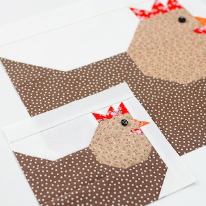 May include: Two brown and white fabric chickens with red and white combs and black eyes. The chickens are sewn together and have a white background.