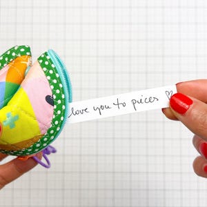 May include: A quilted fabric fortune cookie pouch featuring a colourful geometric pattern in yellow, pink, and blue with a green polka dot border and a light blue zip. A white paper strip extends from the pouch with the text love you to pieces.