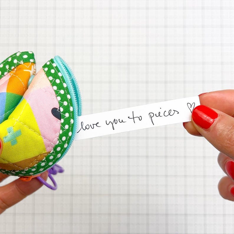 May include: A quilted fabric fortune cookie pouch featuring a colourful geometric pattern in yellow, pink, and blue with a green polka dot border and a light blue zip. A white paper strip extends from the pouch with the text love you to pieces.