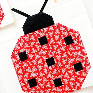 K&ouml;nnte beinhalten: Ein roter Marienk&auml;fer-Patchworkblock mit schwarzen Punkten und einem schwarzen Kopf. Der Marienk&auml;fer ist aus Stoff mit einem kleinen Blumenmuster gefertigt.