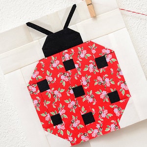 K&ouml;nnte beinhalten: Ein roter und schwarzer Marienk&auml;fer-Patchworkblock mit einem Blumenmuster. Der Marienk&auml;fer besteht aus Stoffquadraten, die zusammengen&auml;ht wurden, um ein geometrisches Muster zu bilden.