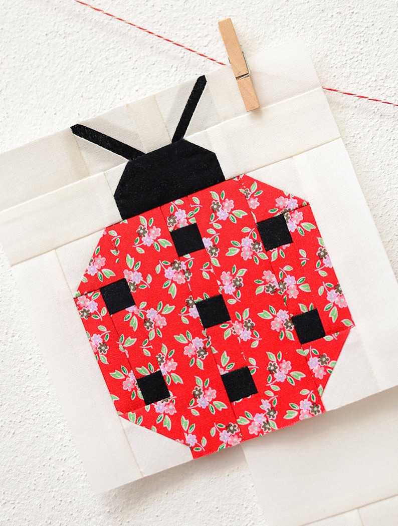 K&ouml;nnte beinhalten: Ein roter und schwarzer Marienk&auml;fer-Patchworkblock mit einem Blumenmuster. Der Marienk&auml;fer besteht aus Stoffquadraten, die zusammengen&auml;ht wurden, um ein geometrisches Muster zu bilden.