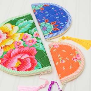 May include: Three semi-circular quilted pouches in green, blue, and orange with floral patterns. The green pouch has a pink tassel, the blue pouch has no tassel, and the orange pouch has a yellow tassel. Each features a zip closure along the curved edge.