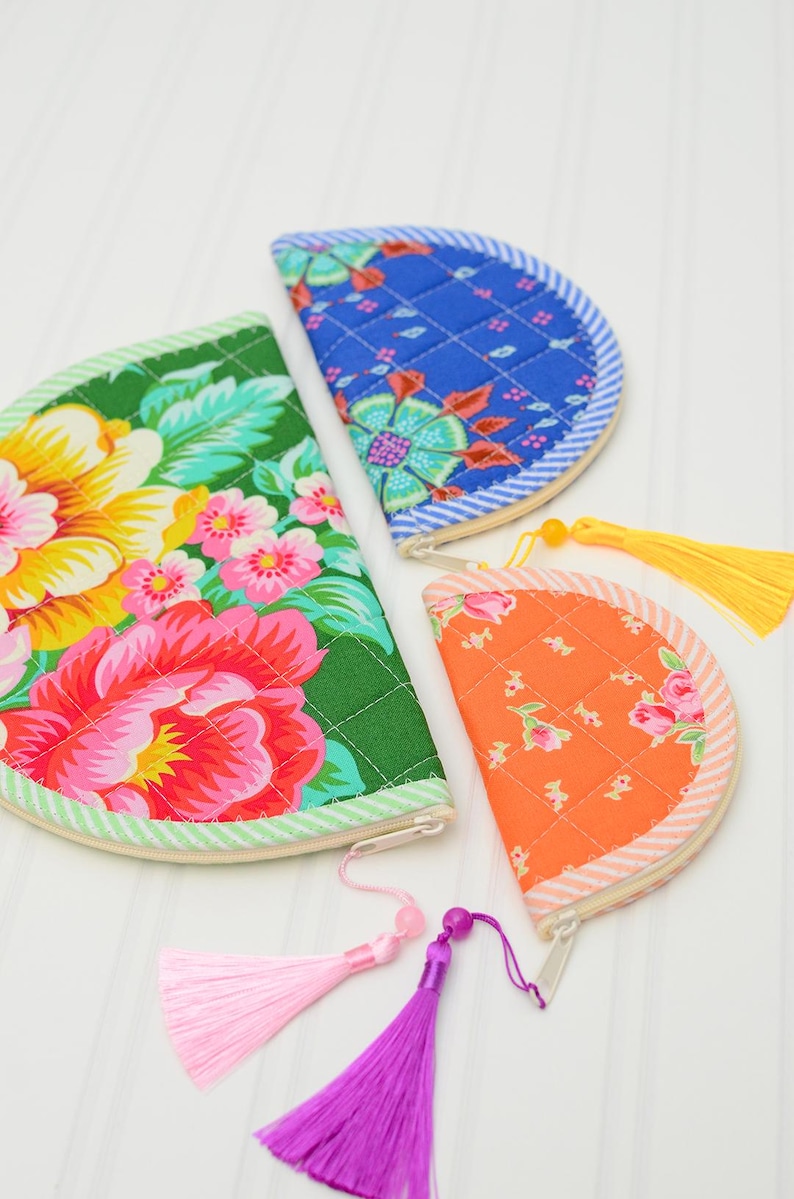 May include: Three semi-circular quilted pouches in green, blue, and orange with floral patterns. The green pouch has a pink tassel, the blue pouch has no tassel, and the orange pouch has a yellow tassel. Each features a zip closure along the curved edge.