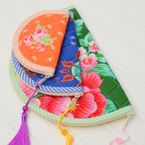 May include: Three semi-circular quilted pouches stacked together. The top pouch is orange with pink roses, the middle is blue with a floral pattern, and the bottom is green with large pink flowers. Each features a zip with a colourful tassel.