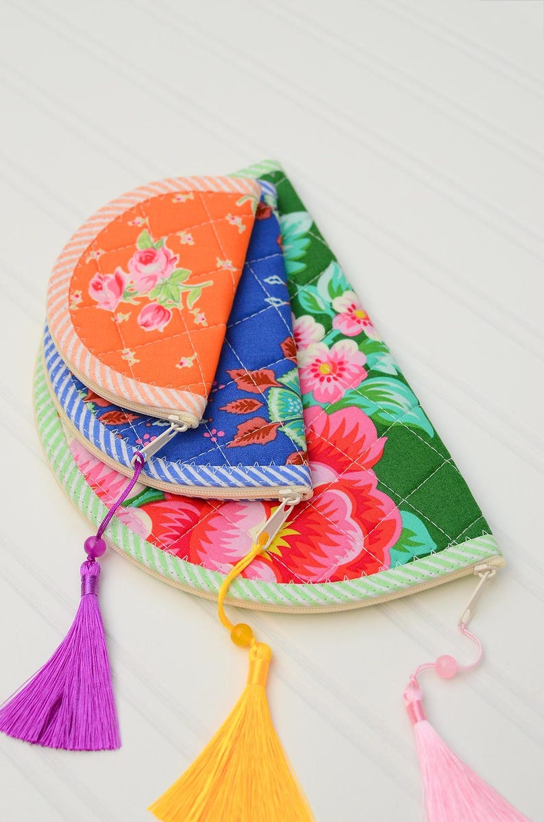 May include: Three semi-circular quilted pouches stacked together. The top pouch is orange with pink roses, the middle is blue with a floral pattern, and the bottom is green with large pink flowers. Each features a zip with a colourful tassel.