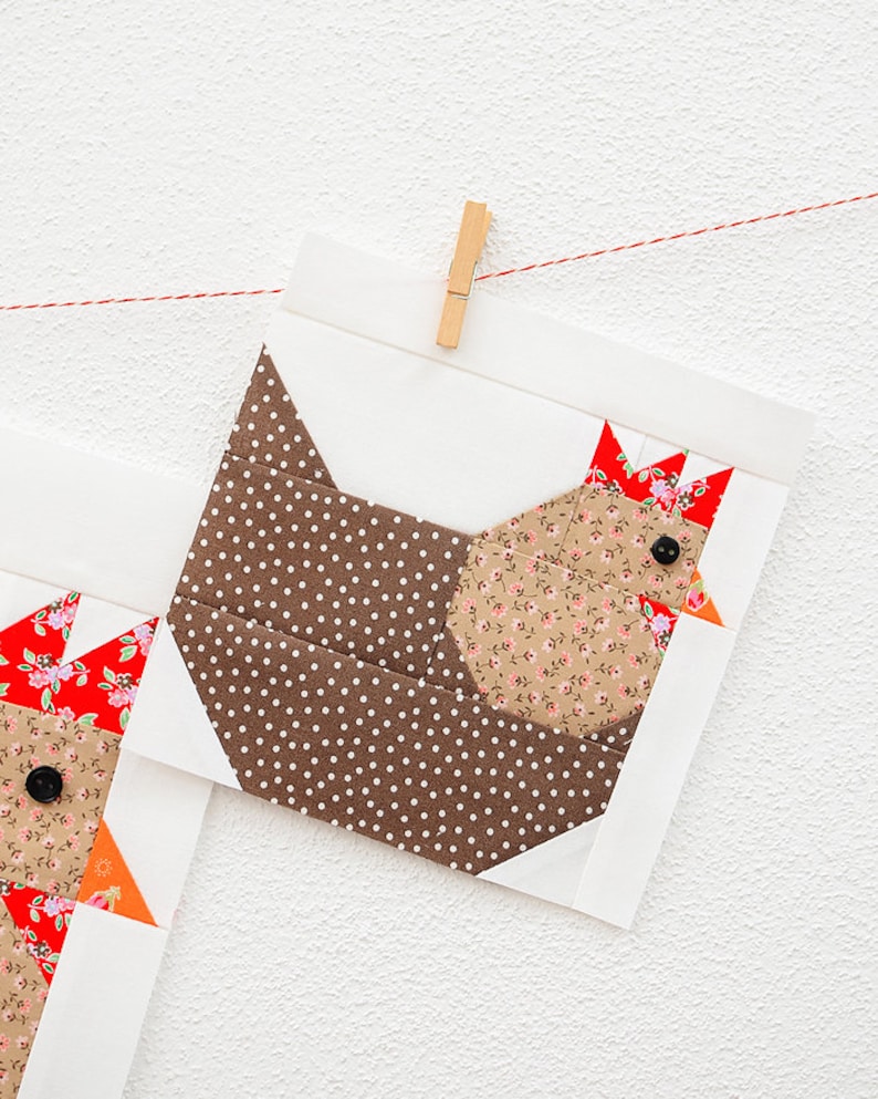May include: A brown and white polka dot fabric chicken applique with a floral print body and red and white floral print comb. The applique is hanging from a red string with a wooden clothespin.