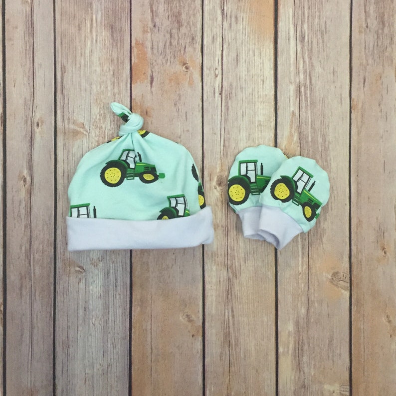 May include: A set of baby accessories featuring a light blue hat and matching mittens. Both items have a repeating pattern of green tractors on a light blue background.