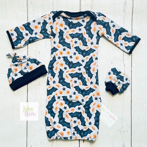 Puede incluir: Un conjunto de beb&eacute; que incluye un vestido, un gorro y manoplas. El conjunto presenta un fondo blanco con un estampado de murci&eacute;lagos turquesa, hojas naranjas y estrellas. Los pu&ntilde;os y el ribete son negros. La marca "Jake and Beth" es visible.