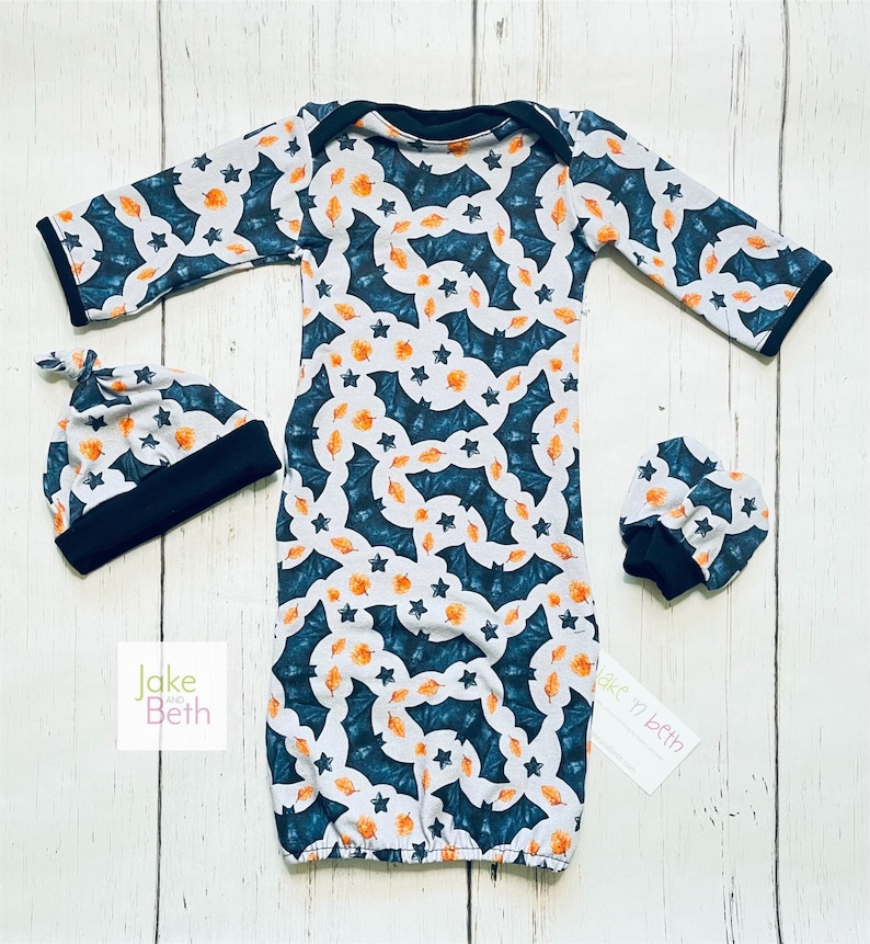 Puede incluir: Un conjunto de beb&eacute; que incluye un vestido, un gorro y manoplas. El conjunto presenta un fondo blanco con un estampado de murci&eacute;lagos turquesa, hojas naranjas y estrellas. Los pu&ntilde;os y el ribete son negros. La marca "Jake and Beth" es visible.