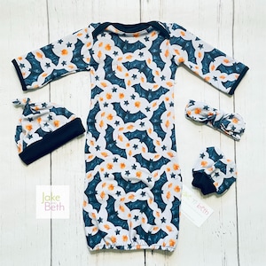 Puede incluir: Un conjunto de ropa para beb&eacute; que incluye un vestido, gorro, diadema y manoplas. El conjunto tiene un fondo blanco con un estampado de murci&eacute;lagos turquesa, hojas naranjas y estrellas. Los bordes del gorro, el vestido y las manoplas est&aacute;n ribeteados en negro.