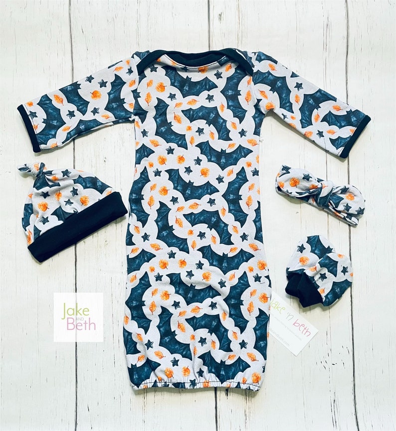 Puede incluir: Un conjunto de ropa para beb&eacute; que incluye un vestido, gorro, diadema y manoplas. El conjunto tiene un fondo blanco con un estampado de murci&eacute;lagos turquesa, hojas naranjas y estrellas. Los bordes del gorro, el vestido y las manoplas est&aacute;n ribeteados en negro.
