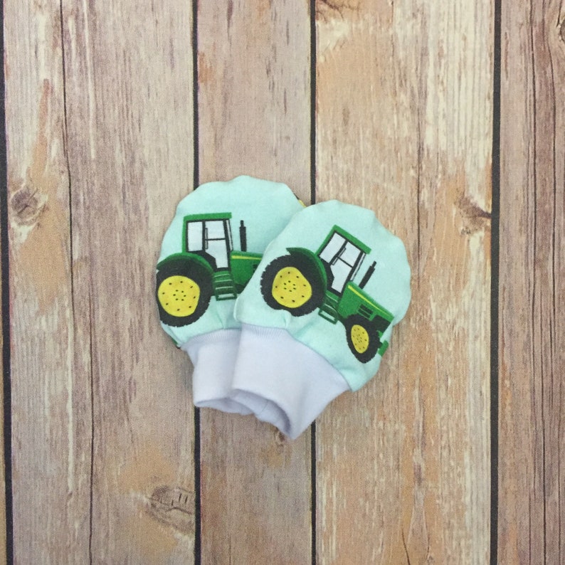 May include: A pair of baby mittens with a green and yellow tractor design on a light blue background. The mittens are made of soft fabric and have a white cuff.