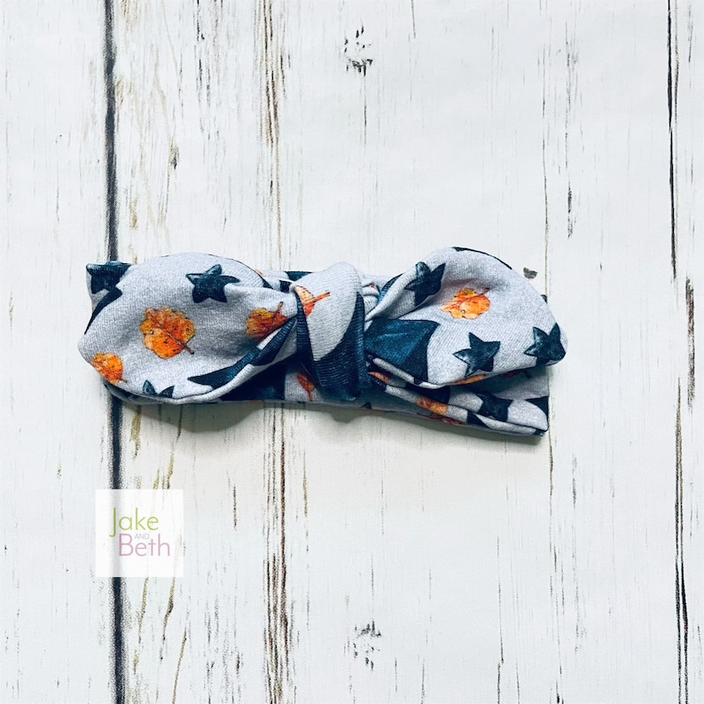 Puede incluir: Una diadema de tela gris con un dise&ntilde;o de lazo anudado. La diadema tiene un estampado de estrellas azul marino y dise&ntilde;os de hojas y bellotas naranjas y marrones. La diadema est&aacute; sobre una superficie de madera blanca.