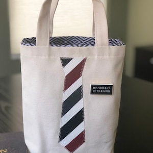 May include: A white canvas tote bag with a black and white striped tie design and the text "Missionary in Training" on a black tag. The bag has a white lining with a black and white geometric pattern.