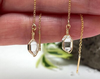 May include: A pair of gold-colored threader earrings. Each earring features a clear, faceted crystal encased in a gold wire cage. The earrings are suspended from delicate gold chains.