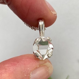 May include: A clear, faceted crystal pendant with a silver-colored bail and chain. The crystal is held in place by a decorative silver setting. The pendant is held between two fingers.
