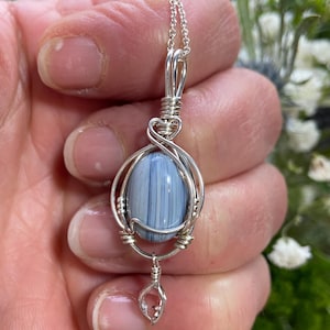 May include: A handcrafted silver pendant necklace featuring a blue and white oval stone. The stone is encased in a wire-wrapped setting, with a small clear crystal charm dangling below. The necklace is held by a silver chain.