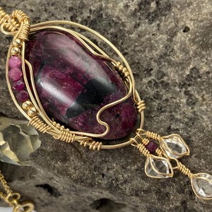 May include: A gold wire wrapped pendant featuring a large, oval-shaped, dark purple and black gemstone. The pendant has a dangling chain with three clear faceted stones and small pink beads.