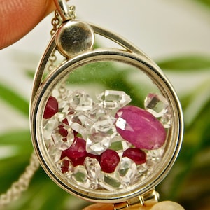 May include: A silver locket pendant with a clear glass insert filled with small red and clear gemstones. The locket is on a silver chain.