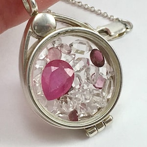May include: A silver locket pendant featuring a circular glass compartment filled with loose, faceted gemstones. The collection includes a prominent pear-shaped pink stone, several smaller clear crystals, and deep red circular stones inside the frame.