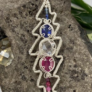 May include: A handcrafted silver pendant necklace featuring a zigzag design. The pendant incorporates a blue gemstone, a clear gemstone, and a pink gemstone. The necklace is displayed on a textured gray surface.