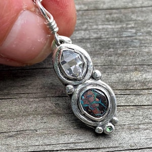 May include: A silver pendant featuring a clear, faceted marquise-shaped stone at the top and a dark, iridescent boulder opal at the bottom. The stones are set in a textured silver frame with small silver spheres and a tiny green accent stone.