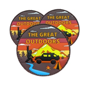 May include: Three brown, round patches with a sunset scene and the text "The Great Outdoors". The patches feature a silhouette of a Bigfoot, a mountain, a car, a tent, and a campfire.