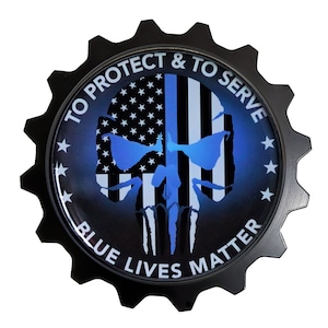 May include: Black metal gear-shaped emblem with a blue and white American flag skull design. The text "To Protect & To Serve" is written around the top of the emblem and "Blue Lives Matter" is written around the bottom.