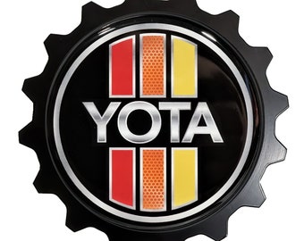 Puede incluir: Emblema de metal negro en forma de engranaje con un logotipo YOTA plateado y rayas rojas, naranjas y amarillas.
