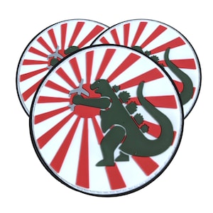 May include: Three round white patches with a red and white rising sun design. A green Godzilla figure is holding a small airplane in its hand. The patches are for sewing or ironing onto clothing.