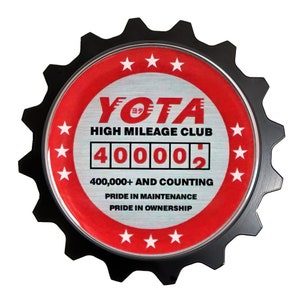 May include: A black and red gear-shaped emblem with the text "YOTA HIGH MILEAGE CLUB" and a digital display reading "400002". The text "400,000+ AND COUNTING" is below the display. The text "PRIDE IN MAINTENANCE PRIDE IN OWNERSHIP" is below the counting text.