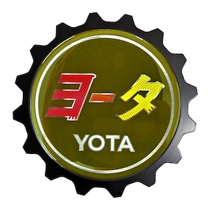 May include: A black gear-shaped emblem with a green background. The emblem features the Japanese characters for "Toyota" in red and yellow, with the word "YOTA" written in white below.